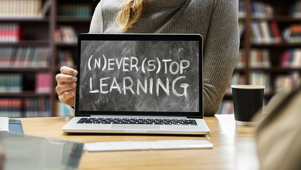 Das Bild zeigt eine Person, die einen Laptop hält. Auf dem Bildschirm steht in Kreideschrift: "(N)EVER(S)TOP LEARNING". Im Hintergrund sind Bücherregale und Bücher zu sehen.